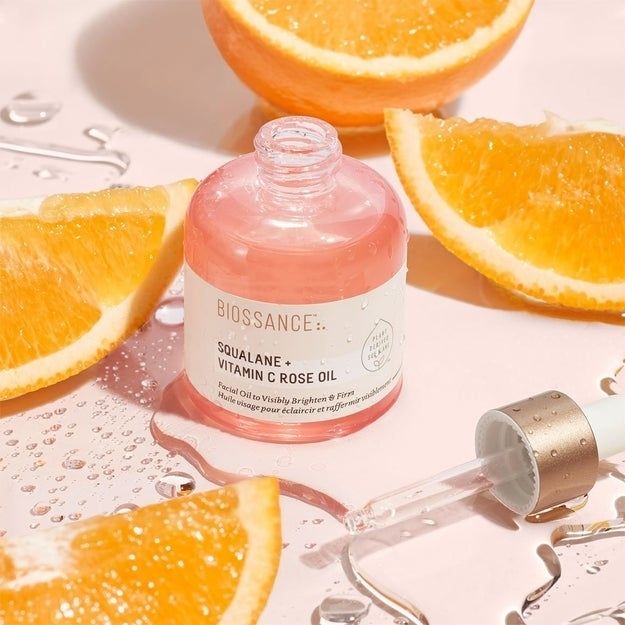 A bottle of Biossance Squalane + Vitamin C Rose Oil, complete with a dropper, is showcased alongside fresh orange slices and water droplets on a glossy surface.