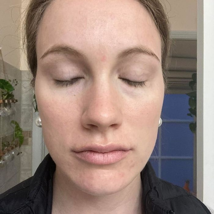 A person with closed eyes and subtle natural makeup is captured indoors, wearing a black jacket over a gray shirt. The focus is on her facial features.