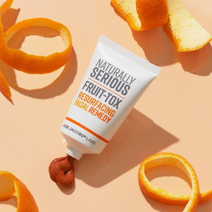 A tube of Naturally Serious Fruit-Tox Resurfacing Facial Remedy is shown alongside scattered orange peels, with a small amount of the product squeezed out for display.