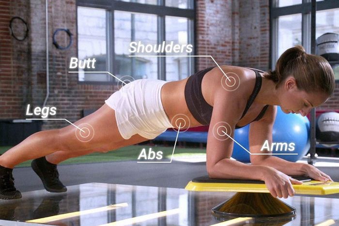 A side view of a person in a plank position while playing the game, with on-screen graphics highlighting the workout their shoulders, glutes, legs, abs, and arms are receiving.