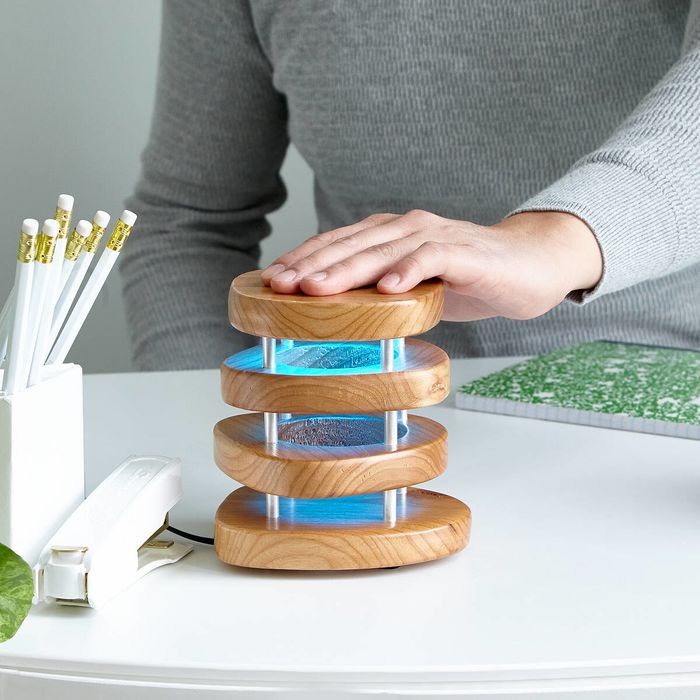 A person places their hand on the long-distance lamp as it emits a soft blue glow. The lamp features four smooth wood pieces connected by metal rods.
