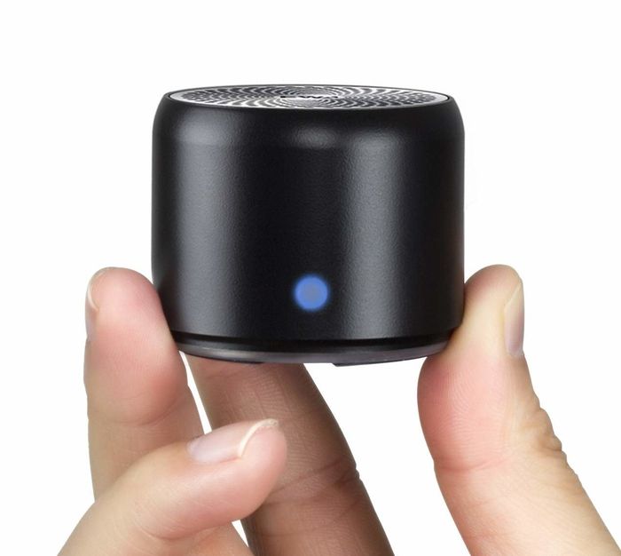 A person holding the small cylindrical speaker between their fingers, showcasing its compact design.