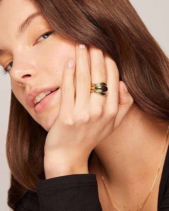 A woman wearing a bold gold statement ring and a delicate necklace, gently resting her hand on her face
