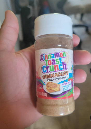 A hand holding a bottle of Cinnamon Toast Crunch Cinnadust seasoning, proudly marked as perfect for toast, cookies, ice cream, and much more