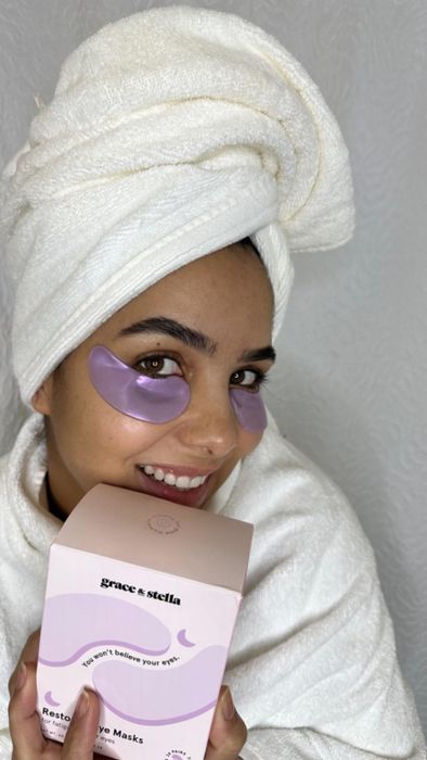 A woman relaxing in a spa environment, wearing purple under-eye masks and a towel on her head, smiling at the camera while holding a Grace & Stella eye mask box