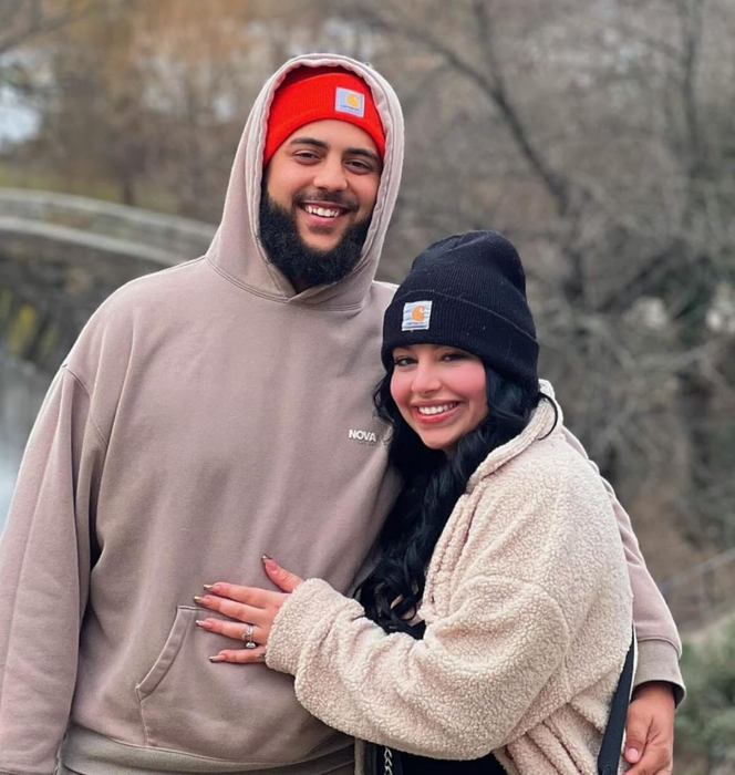 A joyful couple outdoors in winter clothing, both sporting beanies and cozy jackets, sharing a warm embrace in a park-like environment.