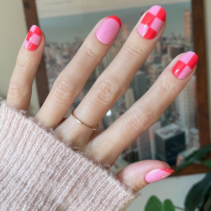 A hand adorned with nail art featuring checkered and striped pink and red patterns, paired with a cozy knit sweater and a simple gold ring