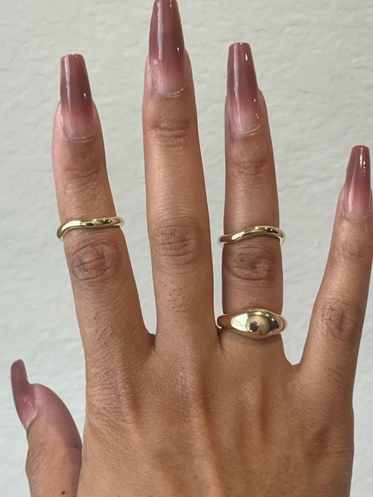 A hand with long, manicured nails showcasing three gold rings: two simple bands and one with a sleek, rounded design