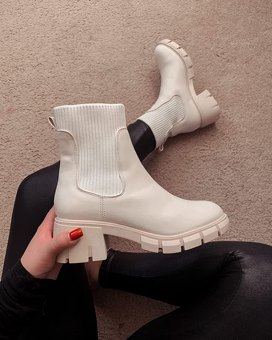 A reviewer is holding one white chunky Chelsea boot in one hand, while wearing the other on their foot.