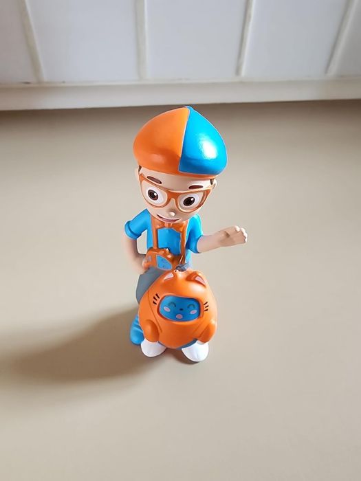 A Blippi toy figure holding an orange bag, wearing colorful glasses and a hat, perfect for children’s gift shopping