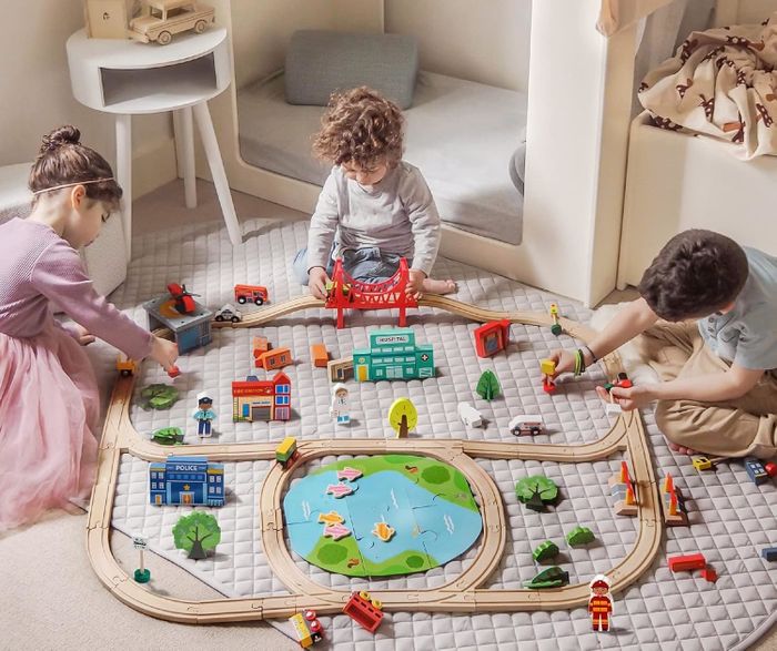 Kids playing with a large train set on a carpet, complete with tracks, vehicles, buildings, and a pond. Text: