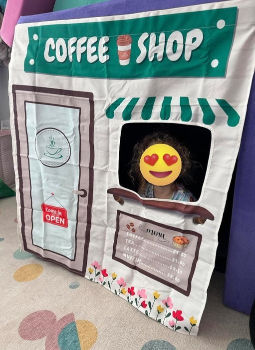 A child playing in a play tent set up like a coffee shop, with a menu listing prices for coffee, tea, lattes, and muffins.