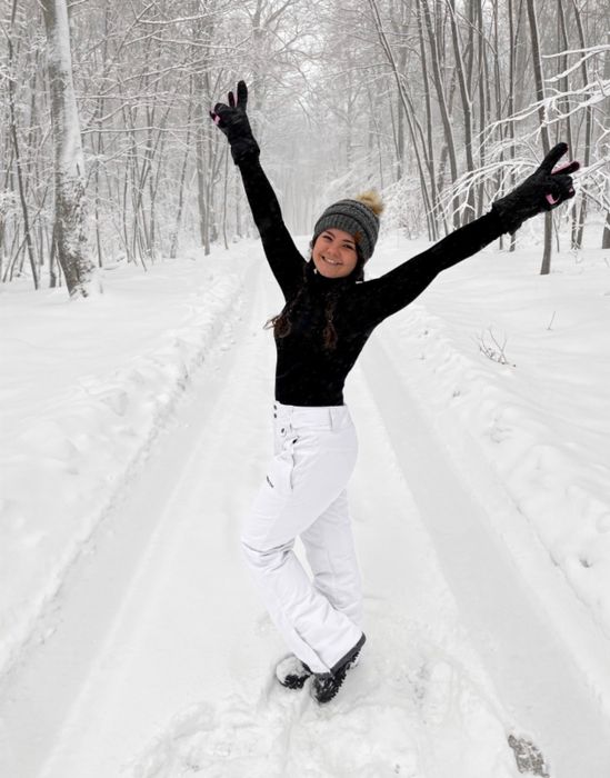 A reviewer wearing a black turtleneck and white pants, standing outdoors in a snowy setting.