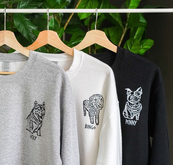 Gray, white, and black sweatshirts featuring dog images, hanging on a clothing rack.