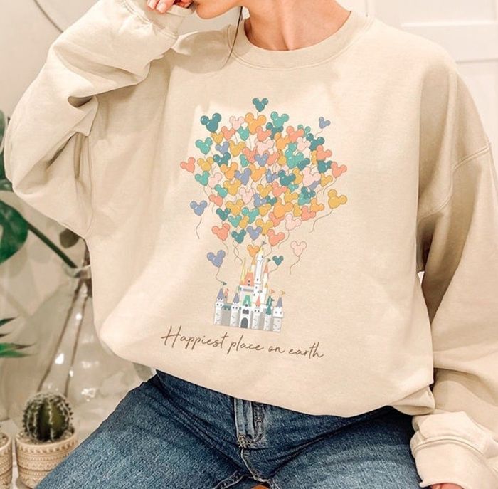 Model wearing a beige sweatshirt featuring an image of Disney’s Magic Kingdom, with Mickey-shaped balloons floating above it and the text: happiest place on earth.