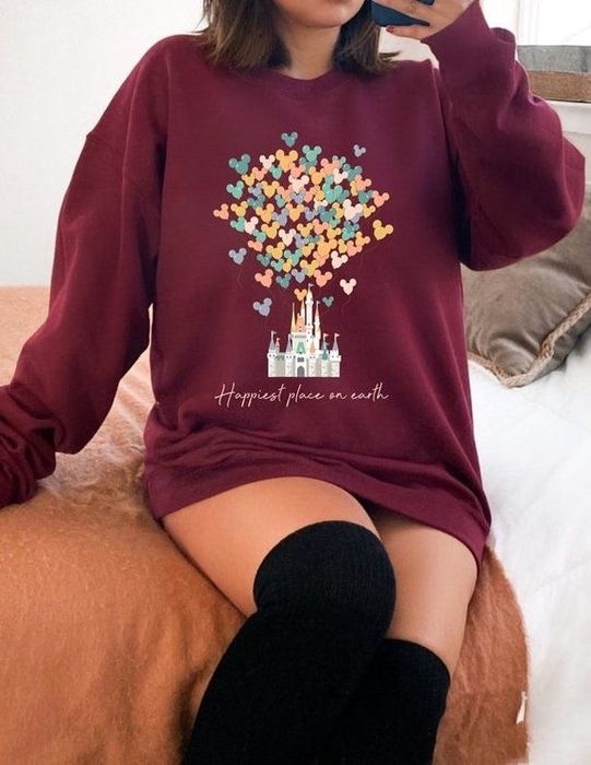 Model wearing a maroon sweatshirt featuring an image of Disney’s Magic Kingdom, with Mickey-shaped balloons floating above it and the text: happiest place on earth.