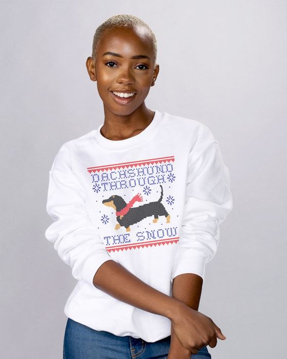 A model is seen in the white crewneck featuring the cross-stitch style text 'dachshund through the snow' and a dachshund sporting a red scarf.