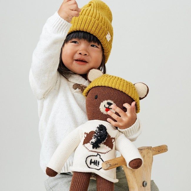 A stuffed brown bear wearing a yellow beanie, sitting next to a child's yellow beanie.