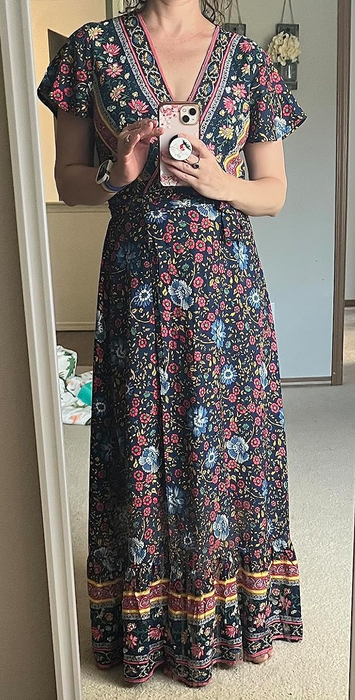 A reviewer is seen standing in front of a mirror, wearing a floral maxi dress with short sleeves, while holding a phone with a decorative case.