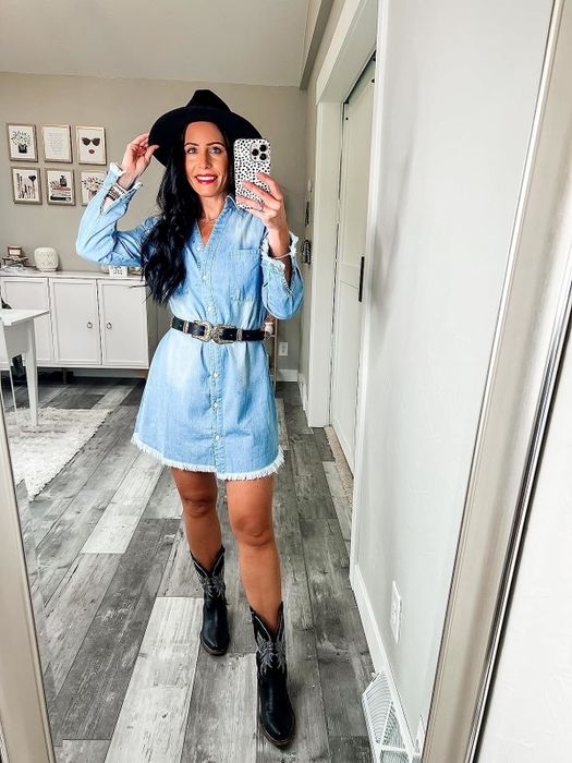 A reviewer dressed in a denim mini dress, paired with a black wide-brimmed hat and black cowboy boots, snaps a mirror selfie in a modern home office space.