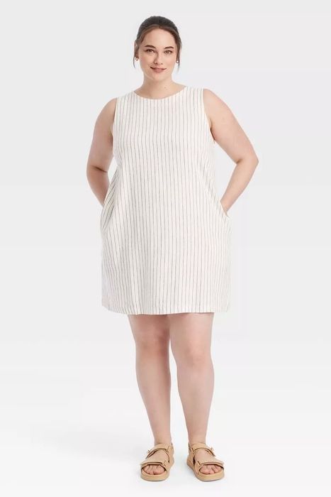 A model wearing a sleeveless, knee-length dress with vertical stripes and hands in pockets, paired with slide sandals and a smile.