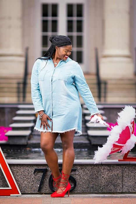 A reviewer dons a denim dress and red shoes, striking a pose outdoors while holding a pink and white feathered umbrella.