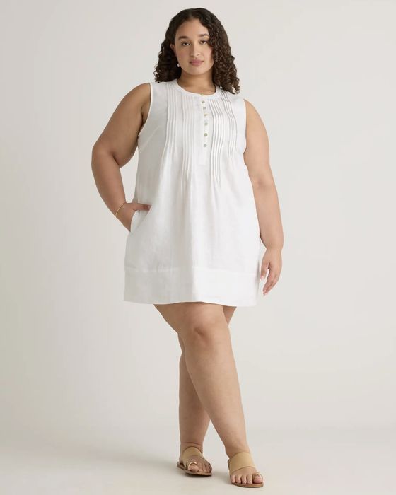 A model wearing a sleeveless, knee-length linen dress with button detailing and sandals.