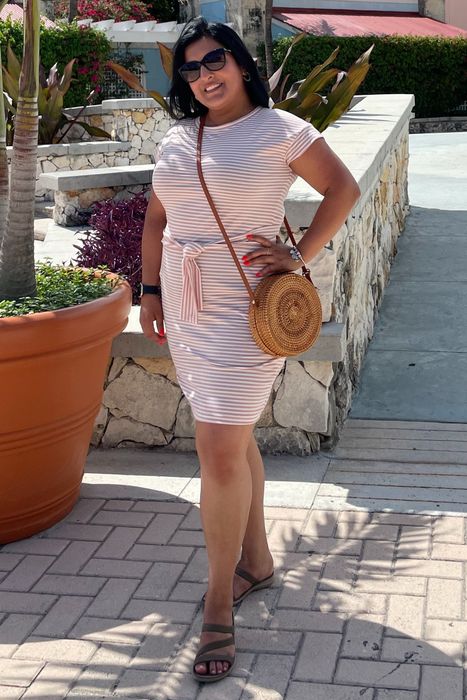 A reviewer steps onto a pathway, wearing a pink and white striped short-sleeve dress and sunglasses, holding a round straw bag, surrounded by potted plants and palm trees.