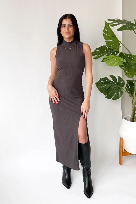 A woman is modeling a sleeveless, form-fitting dress with a side slit, paired with black boots and simple gold jewelry, standing near a potted plant.