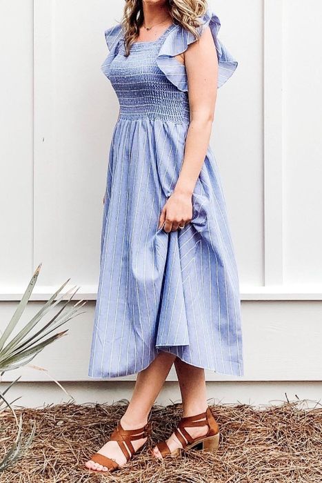 A reviewer models a knee-length, blue striped sleeveless dress with a smocked bodice and a flowing skirt. The background features natural ground, a white wall, and a small plant.