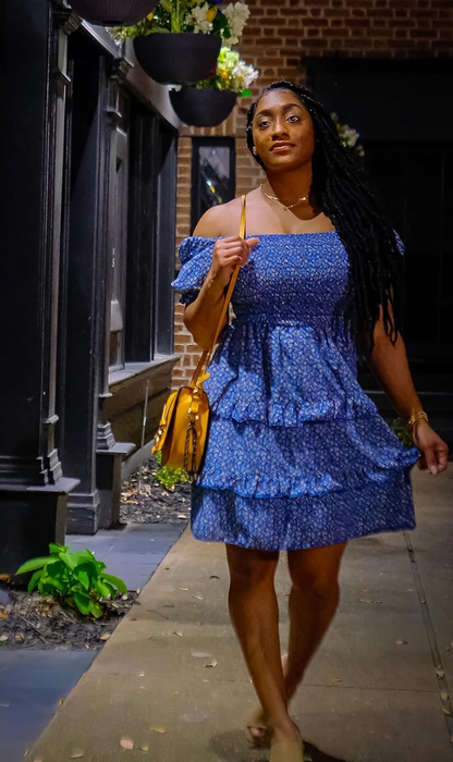 A woman in a blue ruffled dress and a shoulder bag, walking down a city sidewalk at night