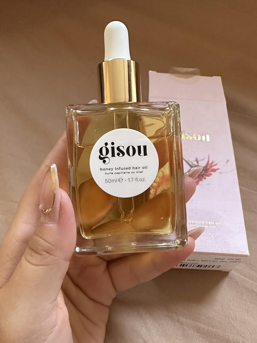A hand holding a 50ml bottle of Gisou honey-infused hair oil (1.7 fl oz), with its packaging box in the background