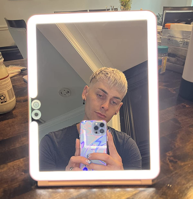 A reviewer taking a selfie in front of a square, illuminated travel mirror.
