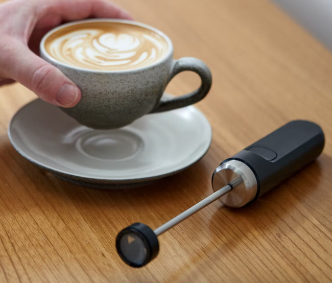A small handheld milk frother beside a beautifully decorated foam latte.