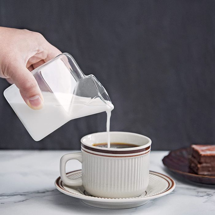 A model pouring milk from a glass container shaped like a milk carton into a coffee cup.