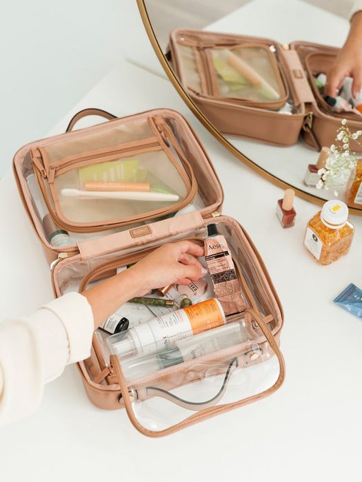 A model is shown filling an open, clear makeup bag with tan edges.