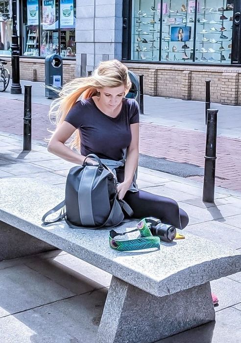A person sitting on a bench, casually wearing a grey backpack with a subtle light grey stripe running down the center.