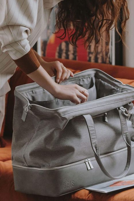 The model demonstrates the duffle bag in grey, showcasing its impressively wide opening.