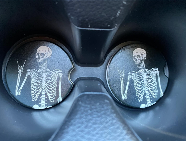Two coasters featuring a skeleton throwing a peace sign at the bottom of a car's drink holder slots.