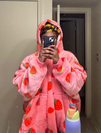 A reviewer takes a mirror selfie while wearing a bright pink, oversized hoodie adorned with red strawberry prints.