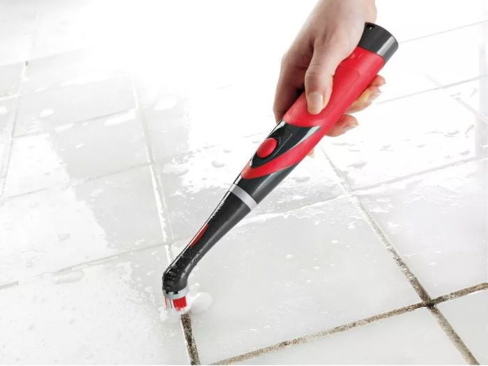 A person using a black and red power scrubber to clean grout between white tiles.