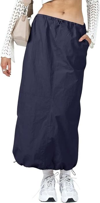 A model is dressed in a white top and navy maxi skirt, accessorized with a beige handbag and white sneakers.