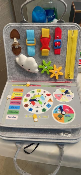 A children's educational busy board featuring zippers, buckles, a clock, and the days of the week for hands-on learning