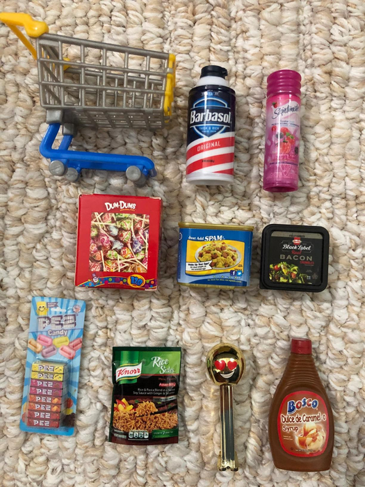 Miniature grocery products including shaving cream, candy, bacon, rice, SPAM, syrup, and a toy shopping cart arranged on a textured surface