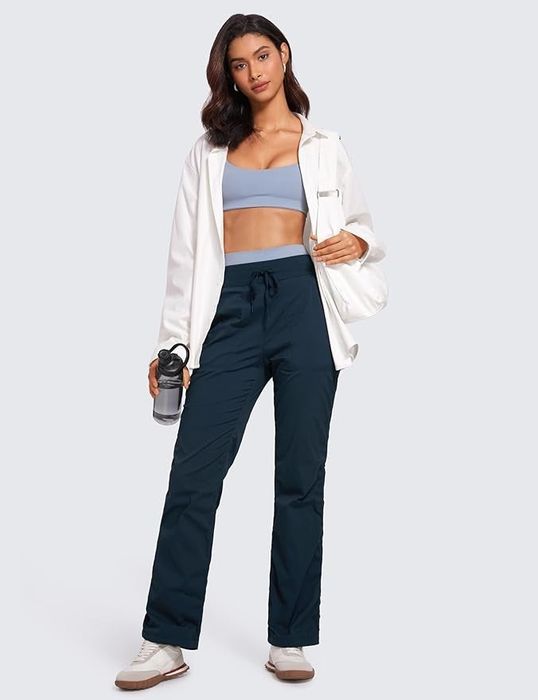 A model dressed in activewear with a light jacket, holding a water bottle, striking a pose for a shopping article.