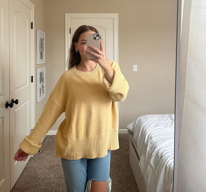 A person takes a mirror selfie, dressed in a casual oversized sweater and ripped jeans, set against a bedroom backdrop