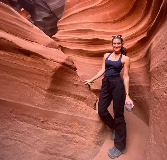 A person stands in a narrow sandstone canyon, wearing a tank top and pants, holding a water bottle and a green item. Sunglasses rest atop their head.