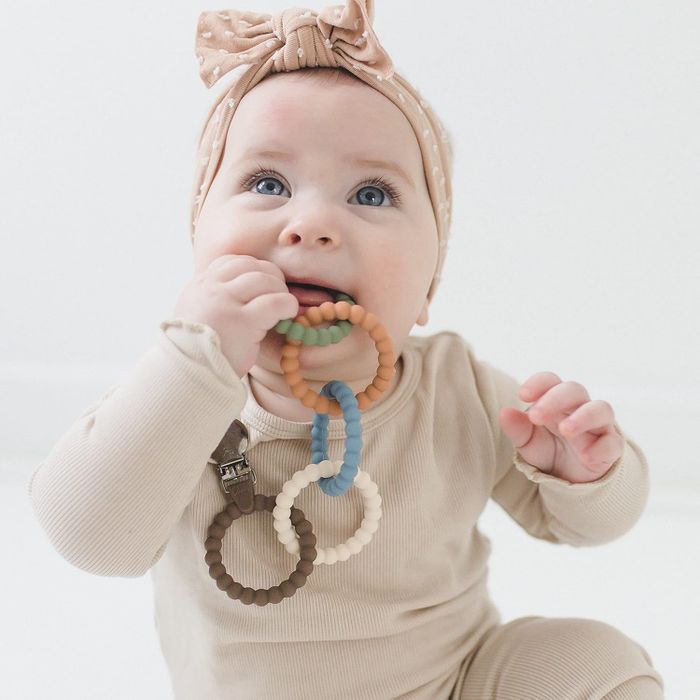 A baby wearing a bow headband plays with a colorful silicone teething ring in a shopping ad