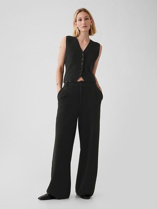 A person stands in wide-leg pants and a buttoned vest, posing with hands in pockets, presenting a minimalist and professional look.