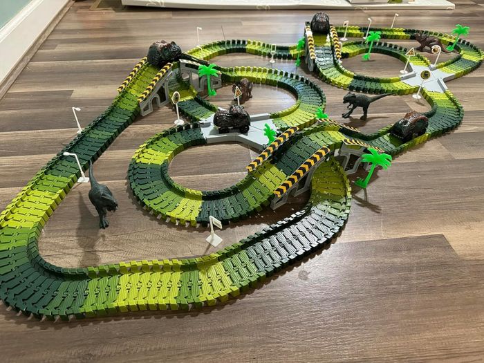 A toy dinosaur race track set with curved paths, a variety of plastic dinosaurs, and palm trees laid out on a wooden surface
